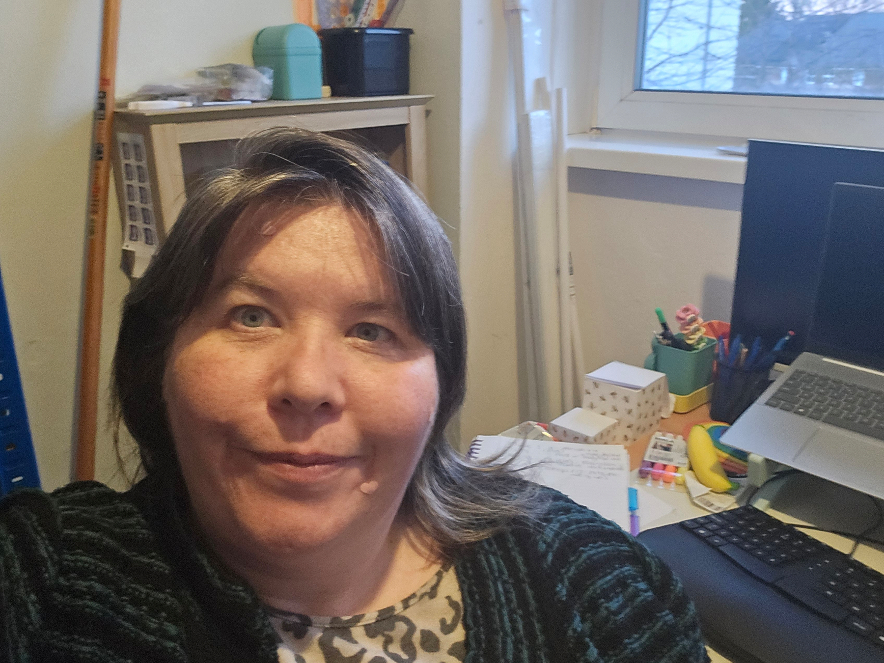 A femme with brown hair is sitting in a home office with a laptop on the desk and various stationery items around.