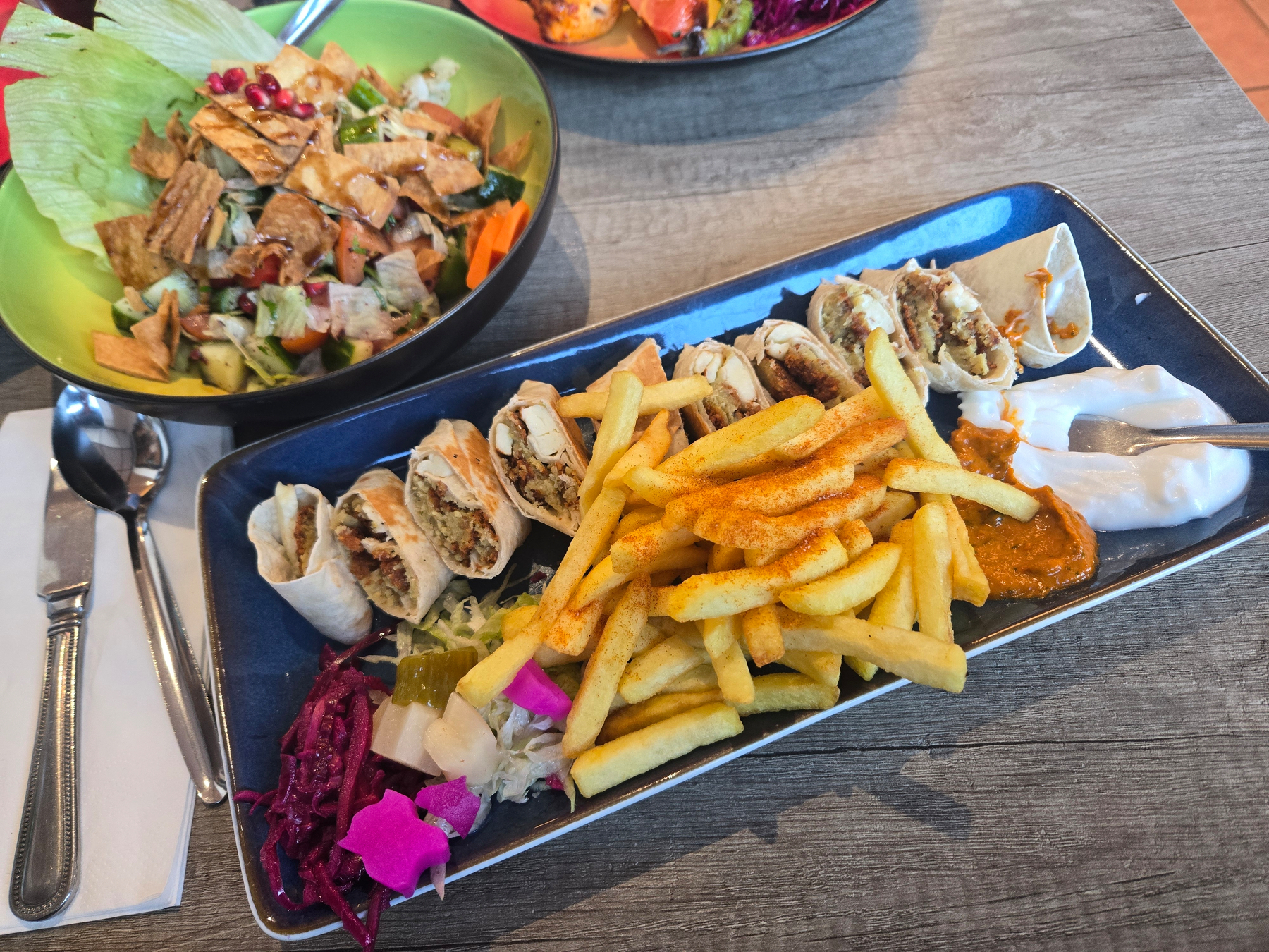 A serving of fries and sliced falafel wraps is accompanied by various sauces and pickles on a dark rectangular plate, with a fattoush salad in the background.